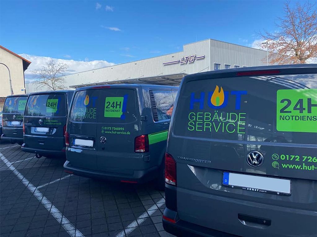 Mehrere dunkelgraue Service-Fahrzeuge mit der Aufschrift HUT Gebäudeservice und 24H Notdienst stehen nebeneinander auf einem Parkplatz vor einem Gewerbegebäude.